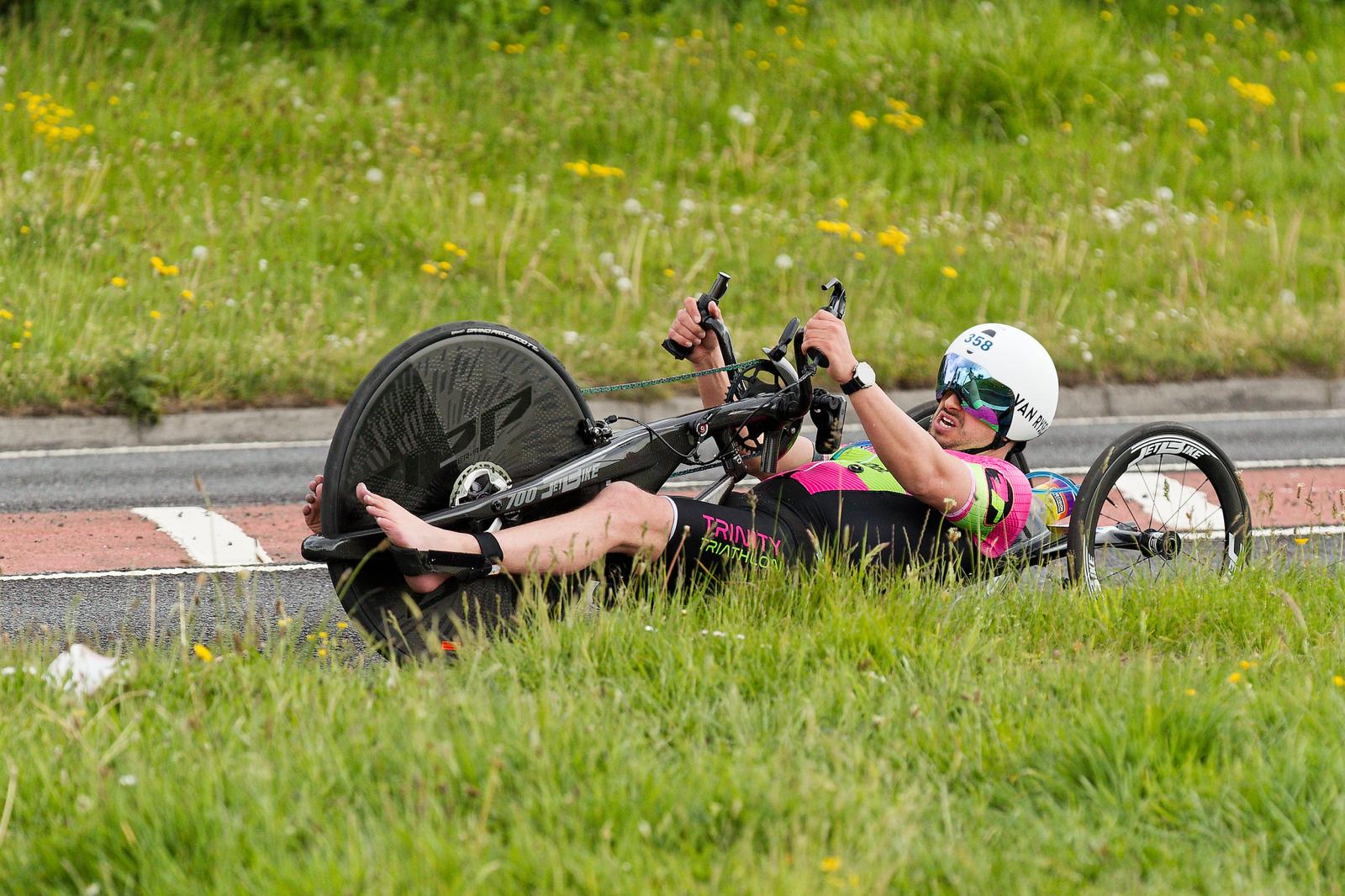 James Bilham racing his handcycle on a closed road, low to the tarmac in Trinity Triathlon kit with spectators lining the verge