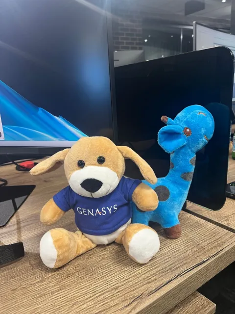 A Genasys-branded toy dog beside a blue toy giraffe on a desk