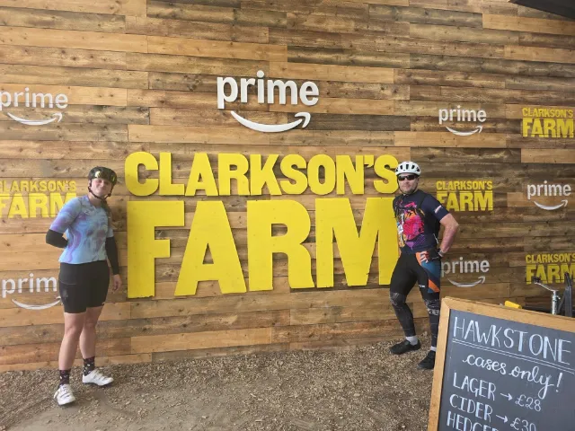 Two colleagues in cycling kit in front of a Clarkson\'s Farm sign